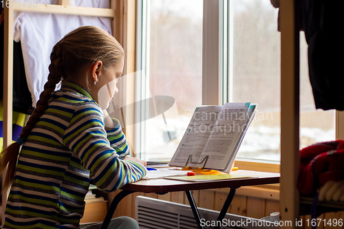 Image of Schoolgirl carefully reads the task in the textbook while doing homework while sitting by the window