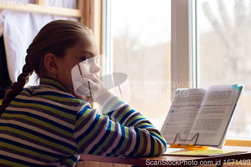 Image of A schoolgirl sits at a table by the window and does her homework, the girl was distracted in thought and looked towards the frame