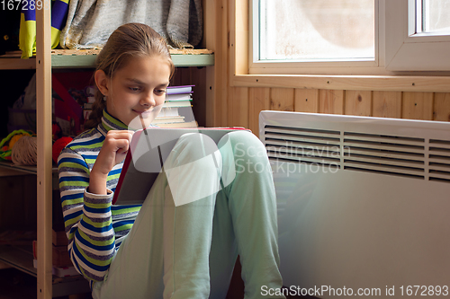 Image of A girl plays in a tablet sitting next to a radiator by a window in a country house