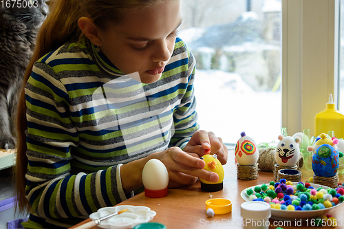 Image of A girl sits at a table by the window and paints Easter eggs with a brush and paints