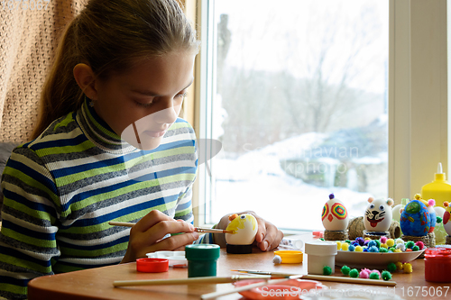 Image of A girl sits at a table by the window and paints Easter eggs with a brush and paints