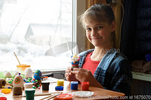 Image of The girl happily looks into the frame while holding a decorated chicken egg craft in her hands