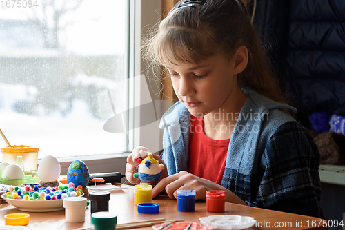 Image of A girl sitting by the window sticks decorative elements on Easter eggs