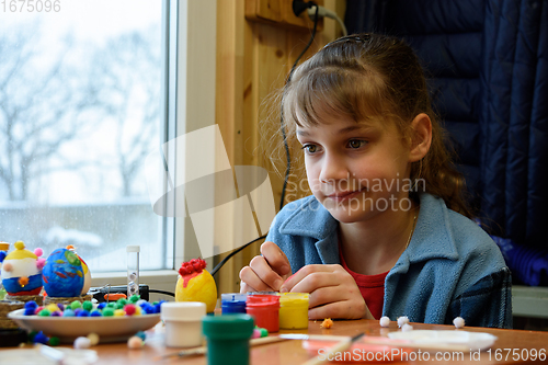 Image of The girl funny thought painting Easter eggs while sitting at the table by the window