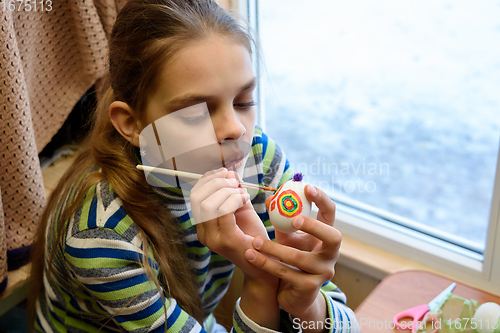 Image of Girl paints Easter eggs while sitting at the table by the window