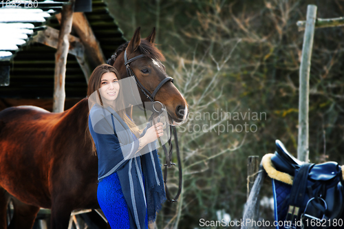 Image of A beautiful girl in a blue dress and a stole stands next to a horse on the background of a forest and wooden buildings