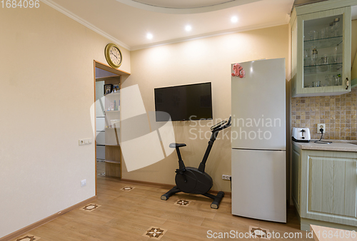 Image of A fragment of the interior of the living room combined with the kitchen, a TV hangs on the wall, an exercise bike and a refrigerator are located nearby