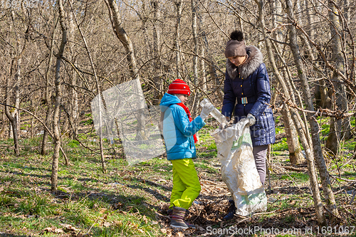 Image of A girl throws a plastic bottle into a trash bag that the girl holds, together they collect garbage in the forest