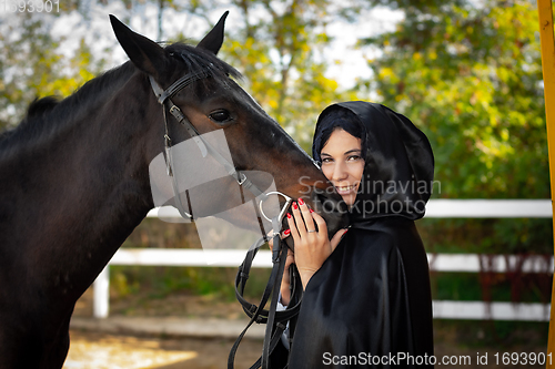 Image of A girl in a black cloak hugs the muzzle of a horse against the background of trees and a fence