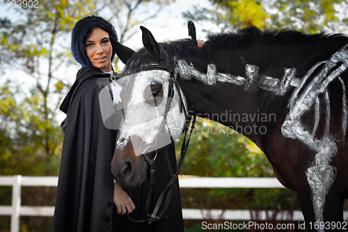 Image of Portrait of a beautiful girl in clothes in a medieval style, next to there is a horse painted with white paint with a painted skeleton