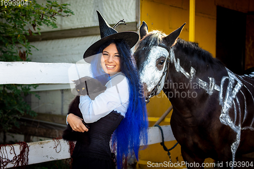 Image of A girl dressed as a witch holds a black cat in her arms and stands by a corral on a farm next to a horse