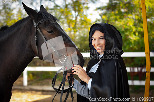 Image of Happy girl in medieval clothes and a black cloak stands with a horse horse on the background of trees and a fence