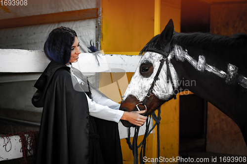 Image of A beautiful girl in medieval style clothes looks at a horse painted with white paint with a painted skeleton