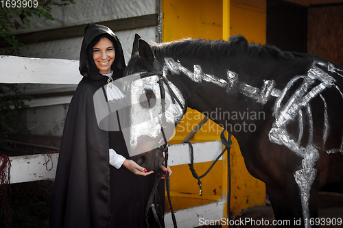 Image of A beautiful girl in medieval style clothes looks happily into the frame and feeds a horse painted with white paint with a painted skeleton