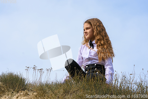 Image of A girl in a white shirt with a bow tie sits on the ground in a field against the background of the sky