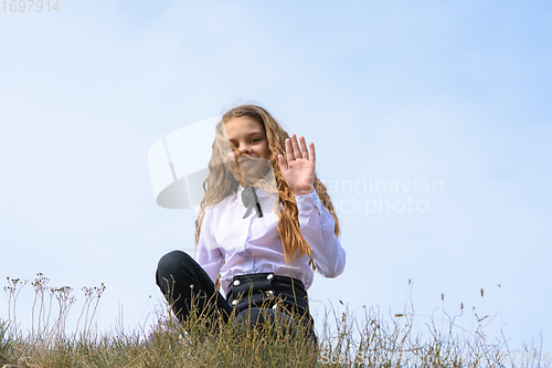 Image of A girl in a white shirt with a bow tie sits on the ground in a field against the background of the sky and waves a pen to the frame