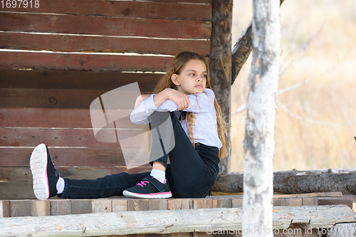 Image of A girl sits on a wooden table against the background of a wall of boards and looks into the distance