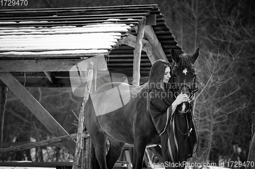 Image of Beautiful girl hugs a horse near wooden buildings on a winter day, black and white photography