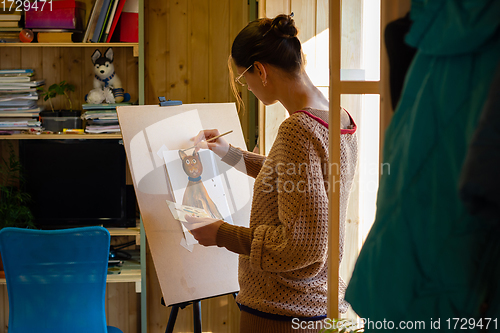 Image of The artist paints a drawing of a cat with acrylic paints on an easel, in a small workshop by the window