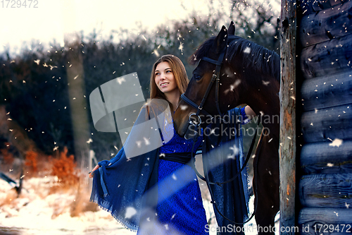 Image of Beautiful girl in a blue stole stands next to a horse near wooden buildings on a snowy winter day