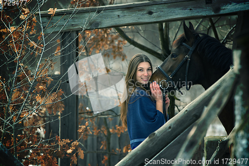 Image of A beautiful girl in a blue stole stands next to a horse on the background of wooden buildings