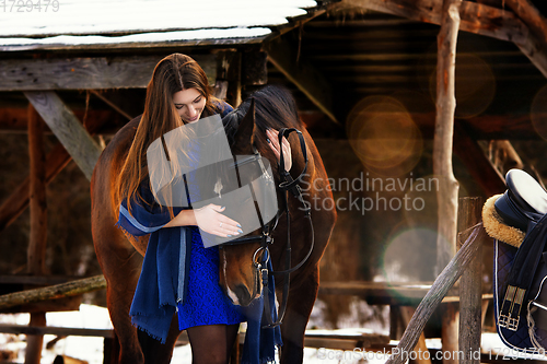 Image of Beautiful girl in a blue stole hugs a horse near wooden buildings on a winter day