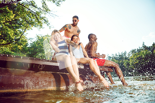 Image of Group of happy friends having fun while sitting and laughting on the pier on river