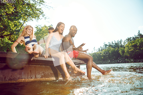 Image of Group of happy friends having fun while sitting and laughting on the pier on river