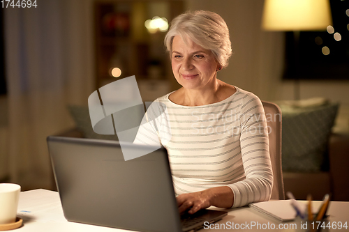 Image of happy senior woman with laptop at home in evening