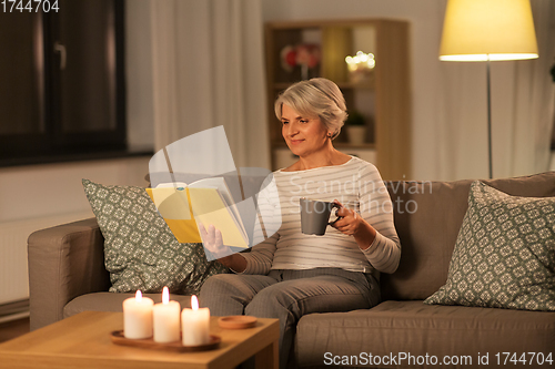 Image of senior woman reading book and drinking tea at home