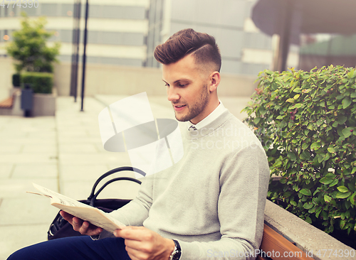 Image of smiling man reading newspaper on city street bench