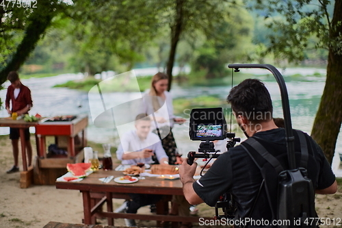 Image of a professionally equipped cameraman is filming a group of people having dinner by the river.