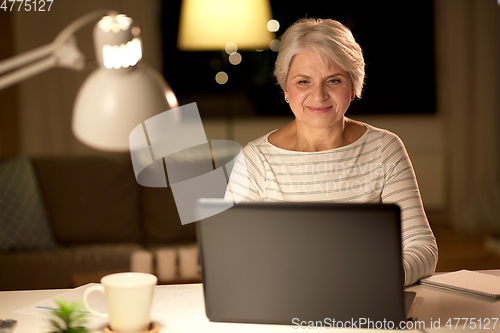 Image of happy senior woman with laptop at home in evening