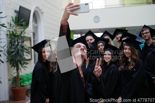 Image of group of happy international students in mortar boards and bachelor gowns with diplomas taking selfie by smartphone