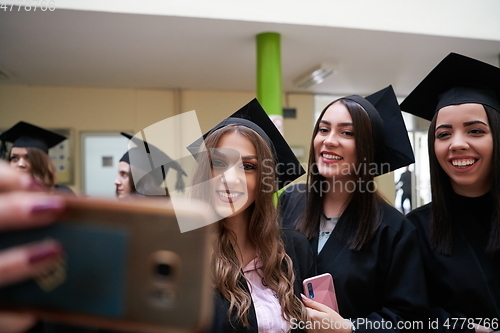 Image of group of happy international students in mortar boards and bachelor gowns with diplomas taking selfie by smartphone