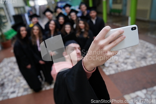 Image of group of happy international students in mortar boards and bachelor gowns with diplomas taking selfie by smartphone