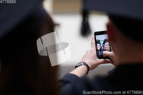 Image of group of happy international students in mortar boards and bachelor gowns with diplomas taking selfie by smartphone