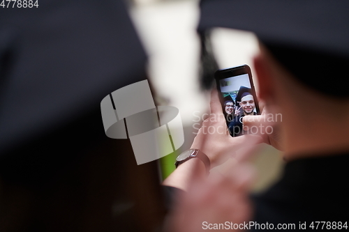 Image of group of happy international students in mortar boards and bachelor gowns with diplomas taking selfie by smartphone