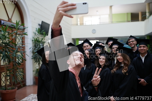 Image of group of happy international students in mortar boards and bachelor gowns with diplomas taking selfie by smartphone