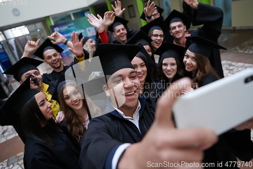 Image of group of happy international students in mortar boards and bachelor gowns with diplomas taking selfie by smartphone