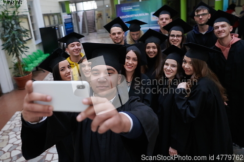 Image of group of happy international students in mortar boards and bachelor gowns with diplomas taking selfie by smartphone