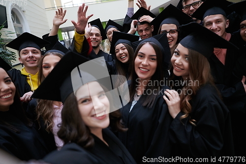 Image of group of happy international students in mortar boards and bachelor gowns with diplomas taking selfie by smartphone