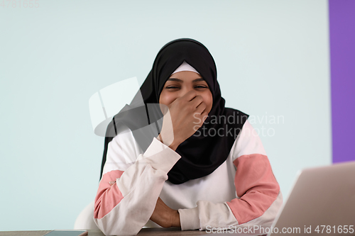 Image of afro muslim woman wearing a hijab sits smiling in her home office and uses a laptop