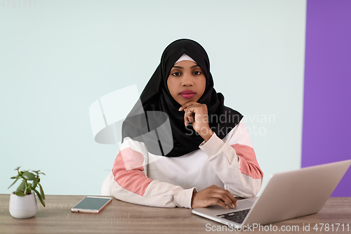 Image of afro girl wearing a hijab thoughtfully sits in her home office and uses a laptop