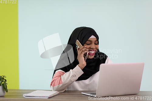 Image of afro muslim woman wearing a hijab sits smiling in her home office and talking on smartphone