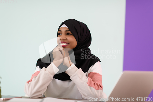 Image of afro muslim woman wearing a hijab sits smiling in her home office and uses a laptop