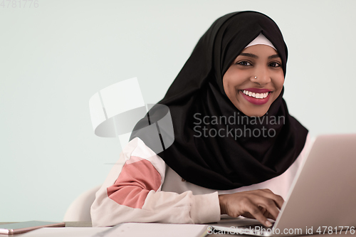 Image of afro muslim woman wearing a hijab sits smiling in her home office and uses a laptop