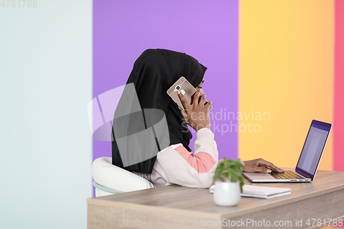 Image of afro muslim woman wearing a hijab sits smiling in her home office and talking on smartphone