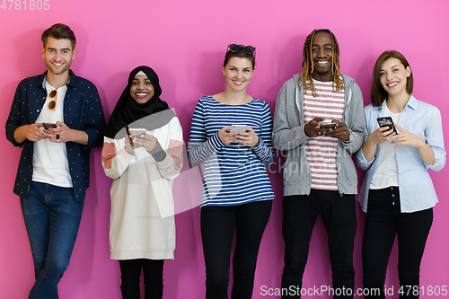 Image of diverse teenagers use mobile devices while posing for a studio photo in front of a pink background