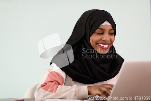 Image of afro muslim woman wearing a hijab sits smiling in her home office and uses a laptop